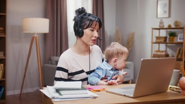 Little boy preventing mother from working. Woman wearing headphones and holding kiddo son. Mother working distantly at maternity leave. Woman taking care of child and working with laptop.