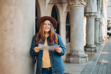Pretty young woman staying on the street holding paper map in hands. 30s girl in hat posing on Market Square in Krakow. Traveling Europe, High quality photo, The Cloth Hall, Vacation concept