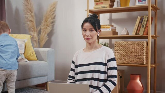 Woman freelancing while on maternity leave. Mother working with laptop and taking care of son. LIttle boy playing behind. Mother looking at camera and smiling. Mom in wireless headphones.