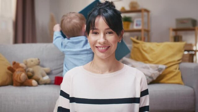 Woman freelancing while on maternity leave. Mother working online and taking care of son. LIttle boy playing with colored cubes. Mother looking at camera and smiling. Joyfull little child.