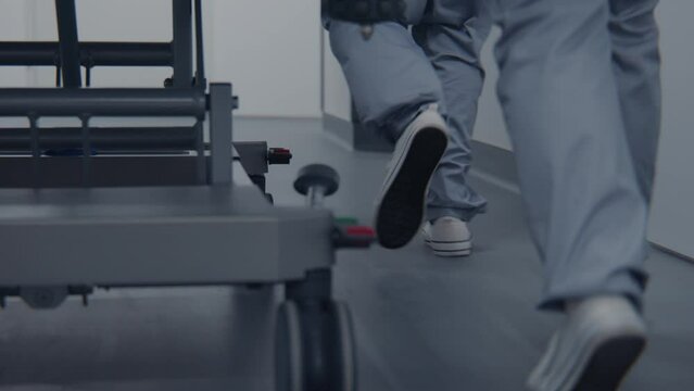 Nurses and paramedics run, push gurney with seriously injured patient to operating room. Medics save life of human. Close up of medical personnel legs. Bright medical facility corridor. Slow motion.