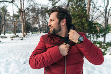 Mid adult man feeling hot while jogging outdoors in winter, unzipping and taking off his red warm...