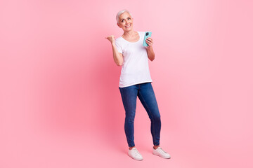 Fototapeta na wymiar Full size photo of cheerful woman wear white t-shirt jeans hold smartphone look directing at empty space promo isolated on pink background