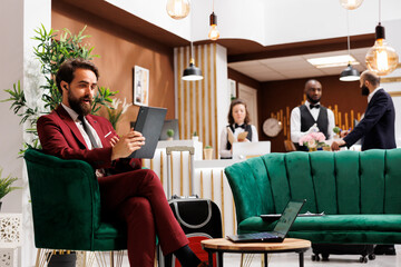 White collar worker talks on videocall, working abroad and travelling. Professional person in suit meeting online with partners to discuss new project ideas, using tablet in lobby at hotel. - obrazy, fototapety, plakaty