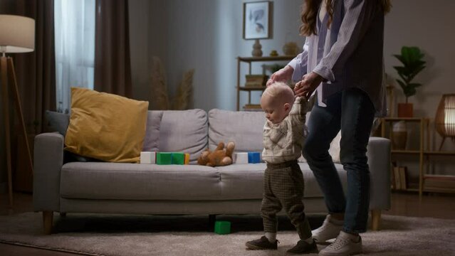 Mother teaching little son to walk. Woman holding baby hands and helping to walk.Baby boy learning how to walk. Mother and toddler walking at living room. Mom spending time with kiddo.