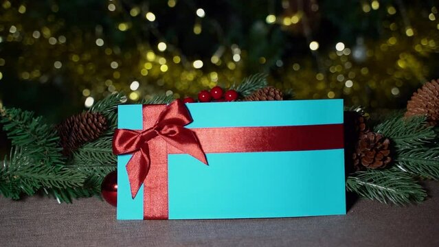 A hand places a blue gift certificate with a red satin ribbon and a bow on a brown tablecloth on a New Year's branch
