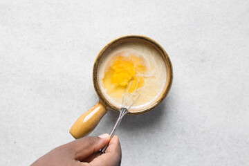 Overhead view of raw egg and milk being whisked in a brown ceramic ramekin, whole raw egg with milk in a ramekin with handle