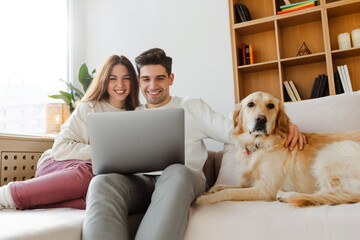 Happy young couple using laptop computer shopping online, watching video sitting with dog on comfortable sofa. Smiling man and woman working from home
