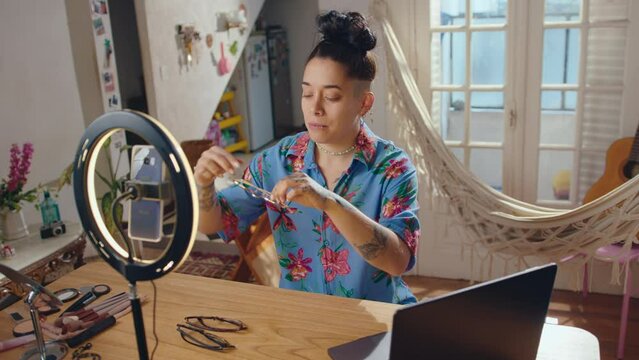 Young female blogger sitting at desk in living room in front of phone and ring light setup, trying on various glasses and talking while filming vlog for social media