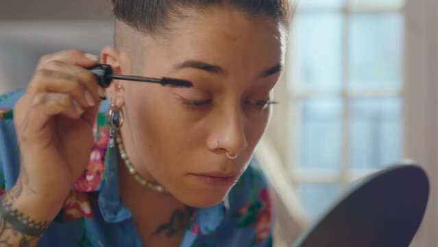 Young girl with tattoos and facial piercing applying mascara and looking in table mirror while doing eye makeup at home