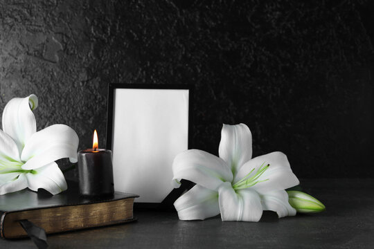 Blank funeral frame, Holy Bible, burning candle and beautiful lily flowers on black background