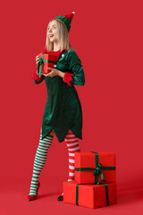 Beautiful young woman in elf costume with Christmas gift boxes on red background