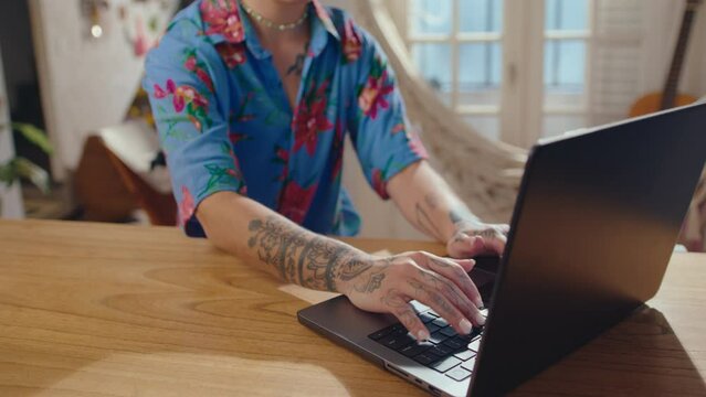 Young girl with undercut hairstyle, face piercing and tattoos sitting at desk in sunlit cozy living room and typing on laptop. Tilt-up shot