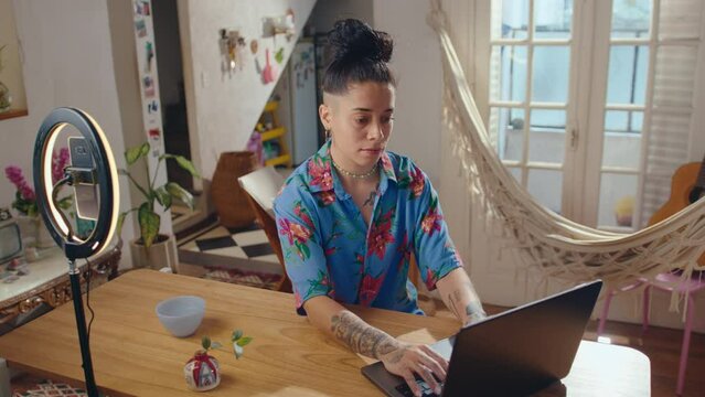 Young female blogger with tattoos and face piercing working on laptop at desk with smartphone and ring light setup on it, creating digital content for social media at home studio