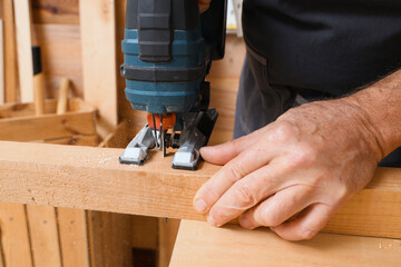 Mature carpenter sawing wooden board with jigsaw at table in workshop, closeup