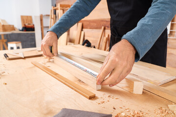 Mature carpenter measuring wooden plank at table in shop, closeup