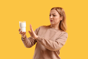 Displeased mature woman rejecting glass of milk on yellow background