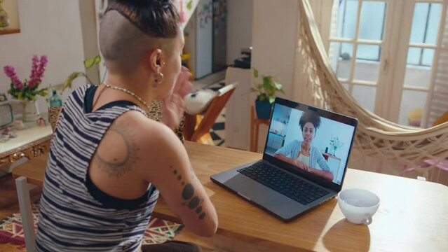 Young girl with tattooed body and undercut hairstyle sitting at table in sunlit living room and chatting with female friend via online video call on laptop