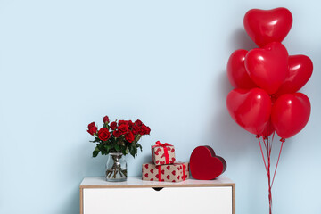 Bouquet of beautiful roses, heart-shaped balloons and gift boxes on commode near blue wall at home...