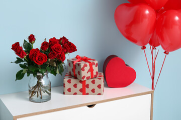 Bouquet of beautiful roses, heart-shaped balloons and gift boxes on commode near blue wall at home...