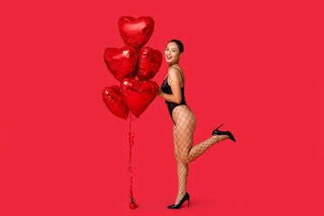 Beautiful young happy Asian woman in sexy underwear with heart-shaped balloons on red background. Valentine's Day celebration - obrazy, fototapety, plakaty