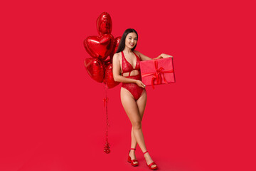 Beautiful young Asian woman in sexy underwear with gift box and heart-shaped balloons on red background. Valentine's Day celebration - obrazy, fototapety, plakaty