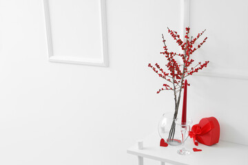 Shelving unit with heart-shaped gift box, candle and red berries near white wall, closeup....