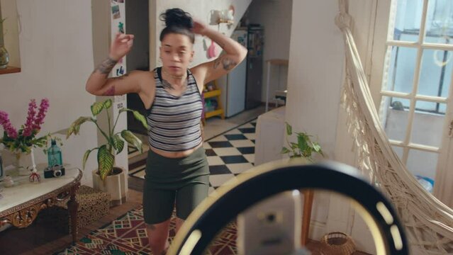 Young female blogger with tattooed body performing dance moves on camera of mobile phone held by tripod with ring light, filming vlog or live streaming on social media at home. Zoom handheld shot