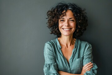 Cheerful middle aged woman with curly hair. Portrait of attractive brunette woman standing with crossed arms and smiling at camera isolated on grey background. Emotion concept - obrazy, fototapety, plakaty