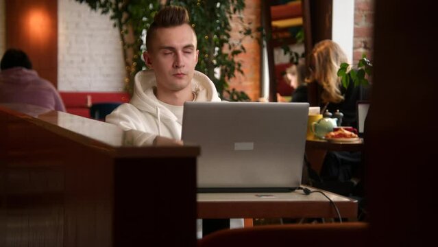 Tired young man is working on laptop in cafe. Stock footage. Handsome young man is writing diploma on laptop in cafe. Young freelancer or student is working on laptop in cafe