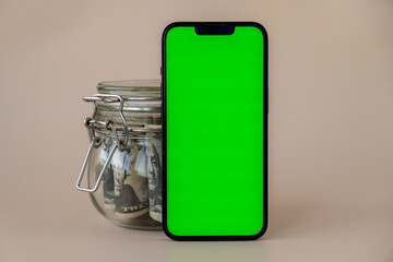 Obraz premium Vertical Green screen on modern mobile phone in background of glass jar full of American currency dollar banknotes on beige background. Cope space for text. Advertisement for application website