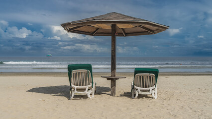Two sun loungers with soft mattresses stand on the beach under a sun umbrella in the shade. The ocean waves are foaming on the shore. Blue sky, clouds. A ship is on the horizon. Malaysia. Borneo.