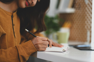 Cropped shot of young woman in yellow sweater writing in journal at her work desk