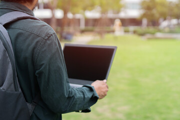 Back view of man standing on grass at park working on laptop. Male wearing backpack and holding laptop