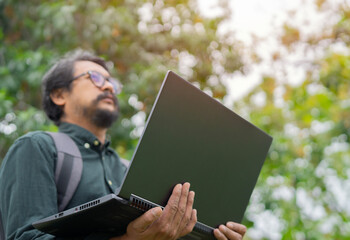 Happy hipster young Asian man wearing glasses standing and working on laptop in the park.
