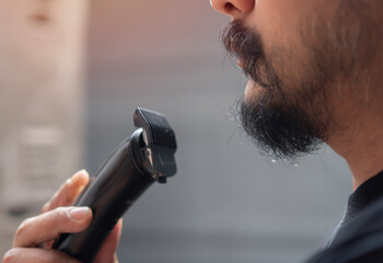 Shaving, grooming and people concept - close up of young man looking to mirror and shaving beard...