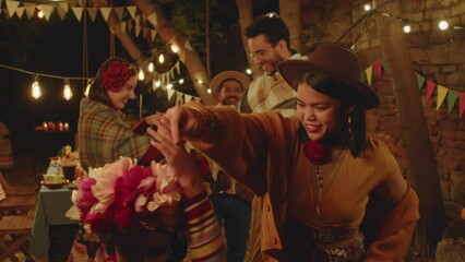 Dolly out shot of cheerful Hispanic friends and family members dancing and having fun together while celebrating Day of the Dead together in backyard at night. One of men playing guitar