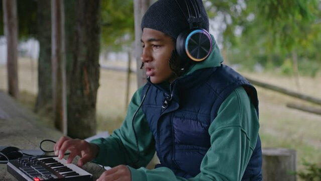 African American teenager wearing headphones plays on MIDI controller outdoors. Young musician, composer creates music, mixes songs, uses laptop for recording while sitting in gazebo in the forest.