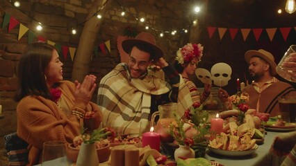 Medium shot of happy Latin family of five having fun during celebration of Day of the Dead outdoors at night, sitting together at festively set dining table decorated with candles and marigold flowers