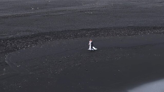 Wedding couple walking on icelandic black sand beach, holding hands, communicating. Rich lava fragments creating dramatic view of vast unique shore. Venue. High quality 4k footage