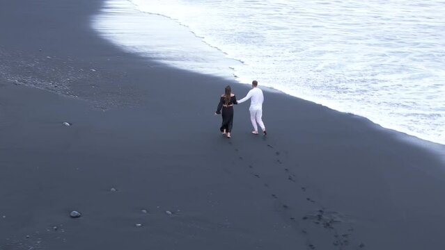 Man in white suit and lady in black long dress running happily on Reynisfjara shore, foaming waves of sea washing their bare feet. Beautiful contrast. High quality 4k footage