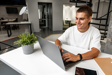 Casual young man in white t-shirt sitting at white desk looking at laptop screen at modern office - Powered by Adobe