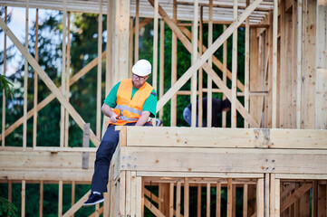 Carpenter constructing two-story wooden frame house near the forest. Bearded man in glasses...