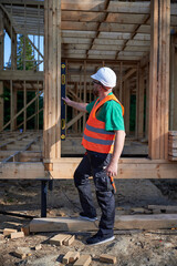 Carpenter constructing two-storey wooden frame house. Man inspects walls for levelness using spirit level, wearing protective overalls, helmet and vest. Concept of modern ecological construction.