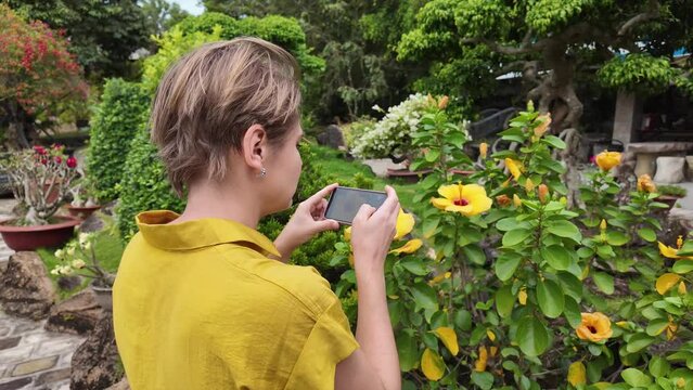 A person in a yellow shirt using a smartphone to take pictures of yellow flowers in a lush garden