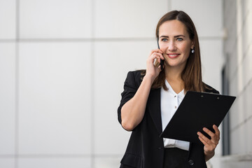 Smiling business-woman in suit looks satisfied with business conversation on phone. Dark-haired lady smiles happily holding documents in hands - obrazy, fototapety, plakaty