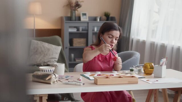 Medium shot of young Caucasian woman with dwarfism wearing nice pink dress sitting at desk in modern living room at daytime and painting with colorful gouache on sheet of paper