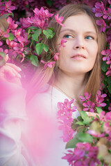 Portrait of beautiful young woman with blonde hair, healthy skin and thoughtful smile wearing casual white blouse or shirt among blooming flowering branches of apple trees with pink lilac flowers