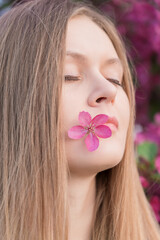 Close up head portrait of beautiful young woman with blonde hair, closed eyes and thoughtful smile with pink elegant flower on lips standing in the garden in front of blooming flowering apple trees