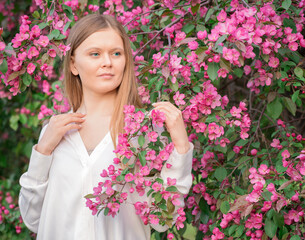 Young attractive woman with blonde hair meditative thoughtful smile wearing casual white blouse or shirt standing among blooming flowering branches of apple tree with pink lilac flowers looking away